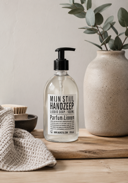 Mijn Stijl handzeep Linnen (500 ml) in een transparante fles met zwart pompje, staand op een robuuste houten plank. Links op de voorgrond ligt een beige, geweven handdoek en daarachter staat een donker schaaltje met een houten afwasborstel. Rechts staat een grote, beige vaas van aardewerk met eucalyptustakken tegen een lichte wand.
