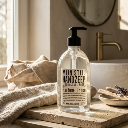 Mijn Stijl handzeep Linnen (500 ml) in een transparante fles met zwart pompje, gepresenteerd op een travertin of natuurstenen plank. Links ligt een lichte handdoek en op de achtergrond is een moderne, gouden kraan met een ronde spiegel zichtbaar bij een wastafel.